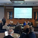 A man holds a microphone next to a screen captioned "Colonial Slave Codes", while a dozen people are seated and looking at the screen